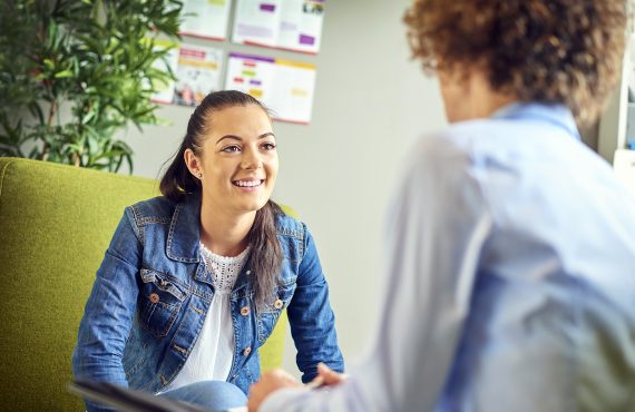 a young woman sits opposite a counsellor and chats happily . They are sitting on armchairs in the counsellor's office . The young woman is smiling as she listens to the advice.