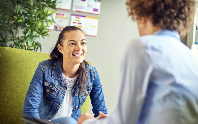 a young woman sits opposite a counsellor and chats happily . They are sitting on armchairs in the counsellor's office . The young woman is smiling as she listens to the advice.