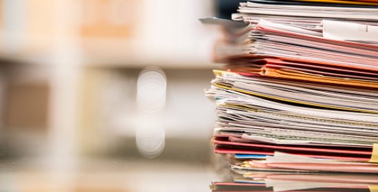 Large stack of file folders, documents, paperwork piled on glass top desk in office.  Bookshelves in background.